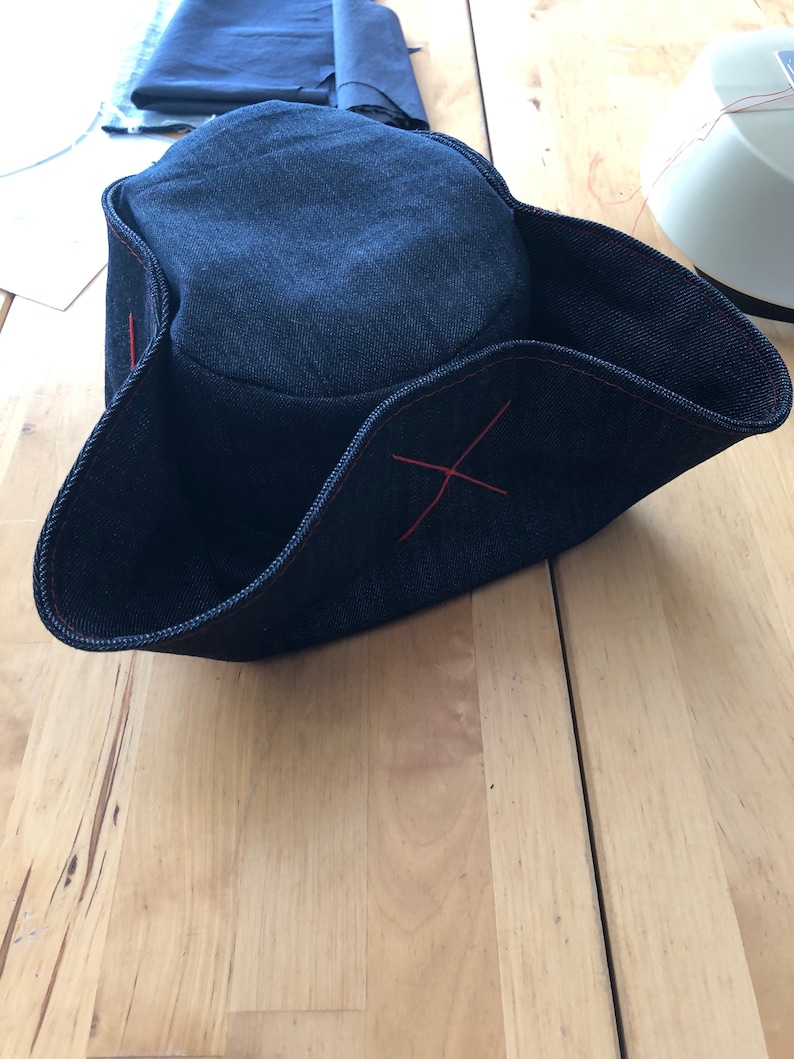 Pode incluir: Um chap&eacute;u tricornio de ganga azul escuro com detalhes de costura vermelha. O chap&eacute;u tem uma aba larga e uma coroa arredondada. A aba &eacute; dobrada para cima em tr&ecirc;s lados. O chap&eacute;u est&aacute; sobre uma superf&iacute;cie de madeira.