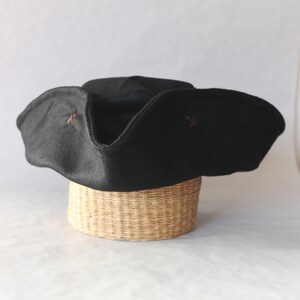 May include: A black tricorn hat with a wide brim and upturned sides, resting on a woven basket. The hat features small, decorative cross-shaped stitching in a contrasting color. The hat is a classic style.