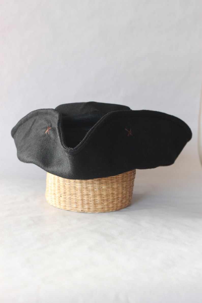 May include: A black tricorn hat with a wide brim and upturned sides, resting on a woven basket. The hat features small, decorative cross-shaped stitching in a contrasting color. The hat is a classic style.