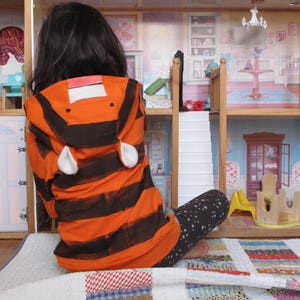 May include: An orange and black striped hooded jacket with white ear accents. The jacket is worn by a person sitting in front of a wooden dollhouse. The dollhouse has multiple rooms with miniature furniture and decor.