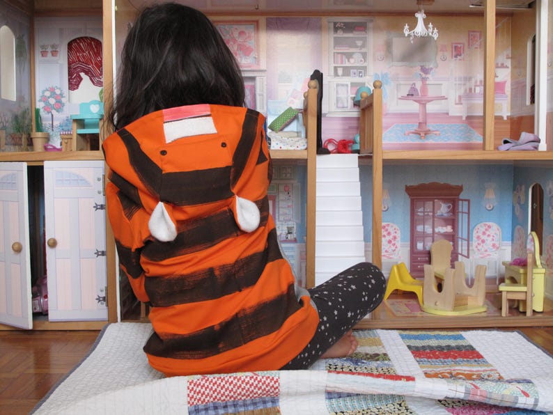 May include: An orange and black striped hooded jacket with white ear accents. The jacket is worn by a person sitting in front of a wooden dollhouse. The dollhouse has multiple rooms with miniature furniture and decor.