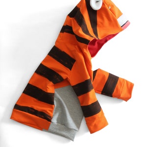 May include: An orange and black striped hooded sweatshirt with a tiger face design. The hood features white ear accents, a pink mouth, and a red interior. The lower portion of the sweatshirt is gray.