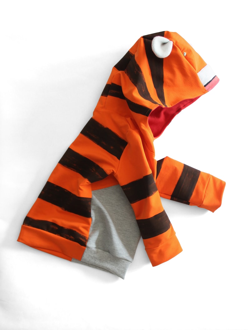 May include: An orange and black striped hooded sweatshirt with a tiger face design. The hood features white ear accents, a pink mouth, and a red interior. The lower portion of the sweatshirt is gray.