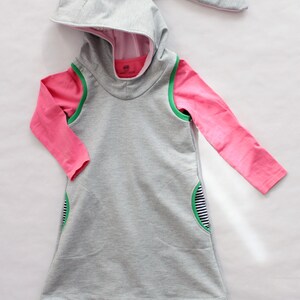 Puede incluir: Un vestido gris con capucha de manga larga y una camisa rosa de manga larga debajo. El vestido tiene ribetes verdes y un bolsillo en el lateral. El vestido se combina con leggings negros.