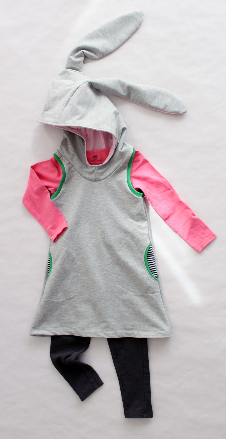 Puede incluir: Un vestido gris con capucha de manga larga y una camisa rosa de manga larga debajo. El vestido tiene ribetes verdes y un bolsillo en el lateral. El vestido se combina con leggings negros.