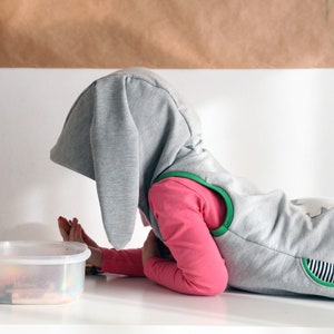 Puede incluir: Un ni&ntilde;o con un disfraz de conejo gris con detalles verdes yace sobre una superficie blanca. El ni&ntilde;o est&aacute; mirando un recipiente de pl&aacute;stico transparente con objetos coloridos en su interior.