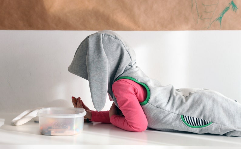 Puede incluir: Un ni&ntilde;o con un disfraz de conejo gris con detalles verdes yace sobre una superficie blanca. El ni&ntilde;o est&aacute; mirando un recipiente de pl&aacute;stico transparente con objetos coloridos en su interior.