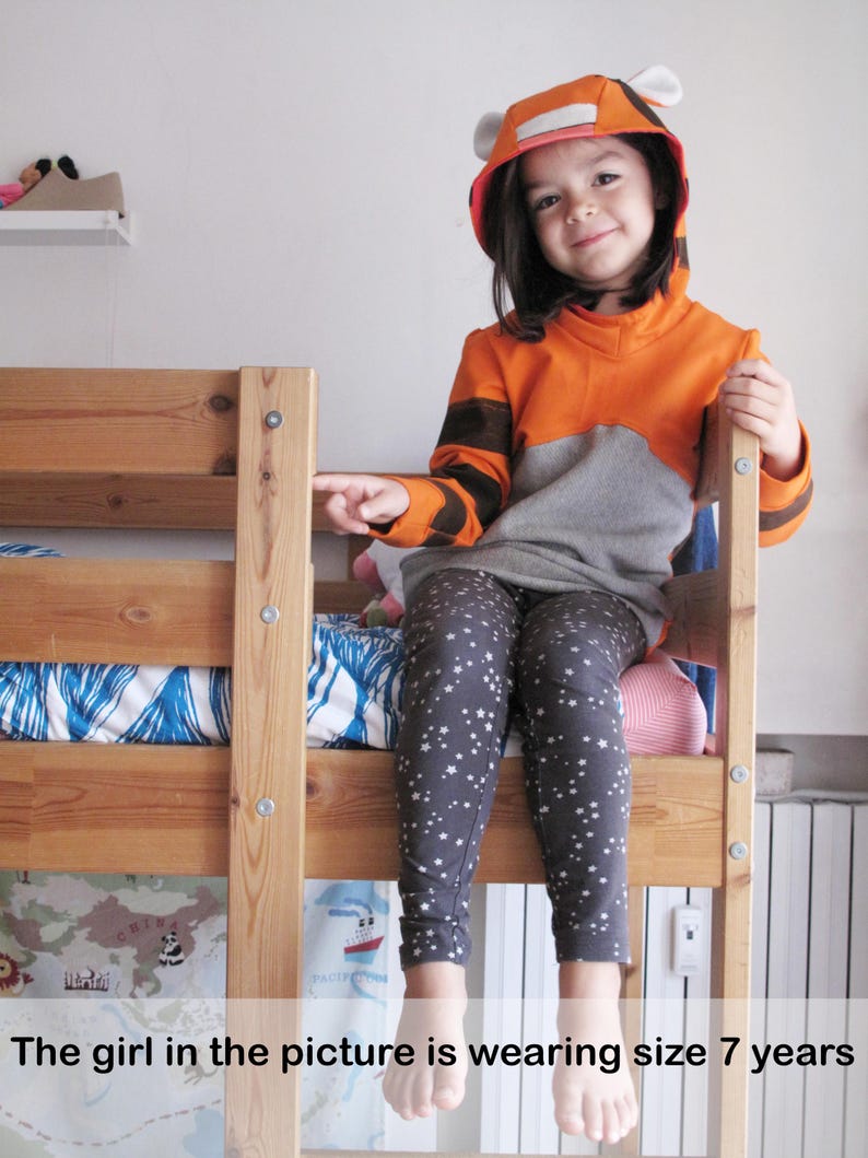 May include: A child wearing a tiger-themed hoodie and star-patterned leggings sits on a wooden bunk bed. The hoodie is orange and gray with black stripes and white ear details. The text on the image says "The girl in the picture is wearing size 7 years."