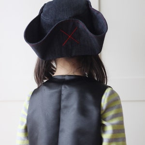 Pode incluir: Um chap&eacute;u de pirata azul escuro com uma cruz vermelha costurada na frente. O chap&eacute;u &eacute; usado com um colete preto e uma camisa listrada. A fantasia de pirata &eacute; adequada para uma crian&ccedil;a.