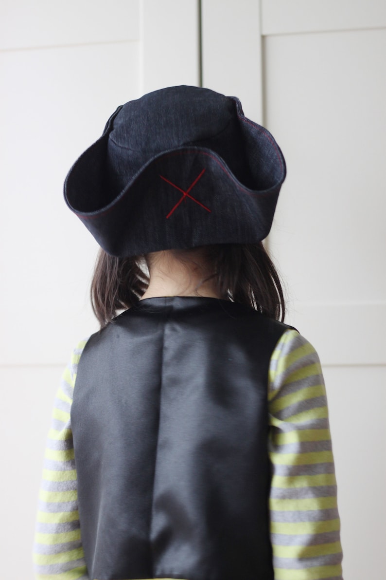 Pode incluir: Um chap&eacute;u de pirata azul escuro com uma cruz vermelha costurada na frente. O chap&eacute;u &eacute; usado com um colete preto e uma camisa listrada. A fantasia de pirata &eacute; adequada para uma crian&ccedil;a.