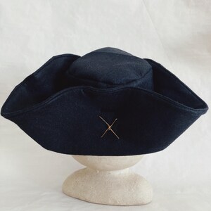 May include: A dark navy blue hat with a wide brim and a small, rounded crown. The hat features a decorative gold-colored cross stitched on the front. The hat is displayed on a cream-colored, rounded pedestal against a white background.