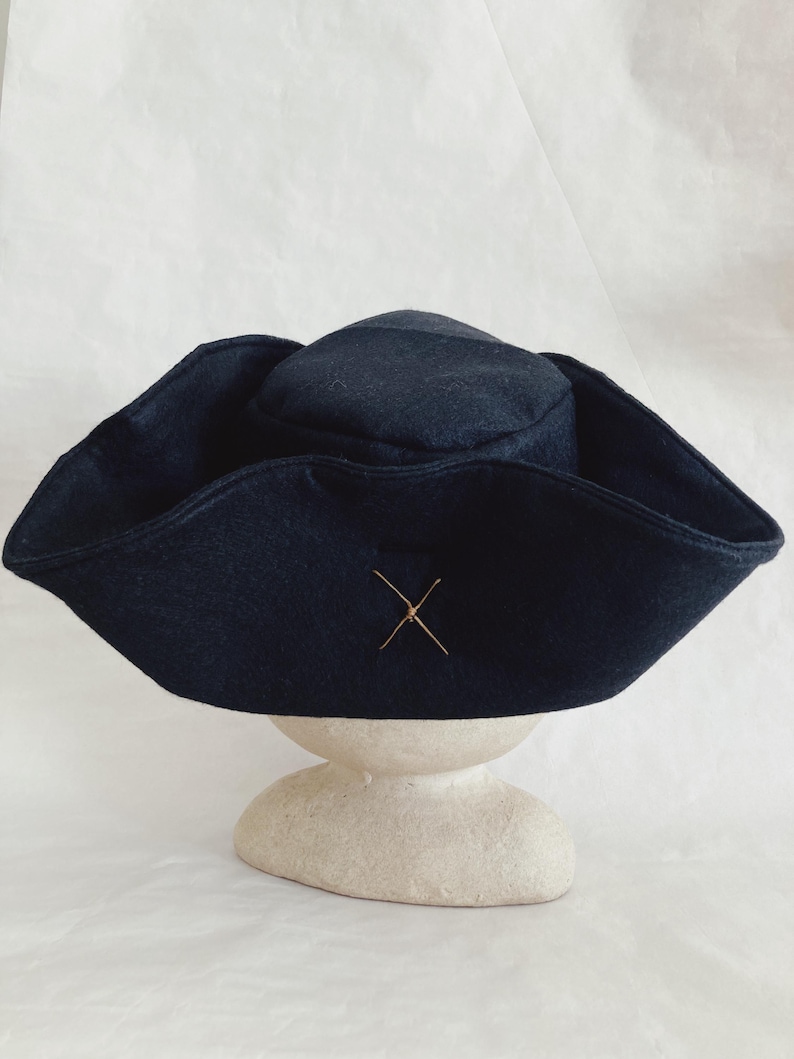 May include: A dark navy blue hat with a wide brim and a small, rounded crown. The hat features a decorative gold-colored cross stitched on the front. The hat is displayed on a cream-colored, rounded pedestal against a white background.