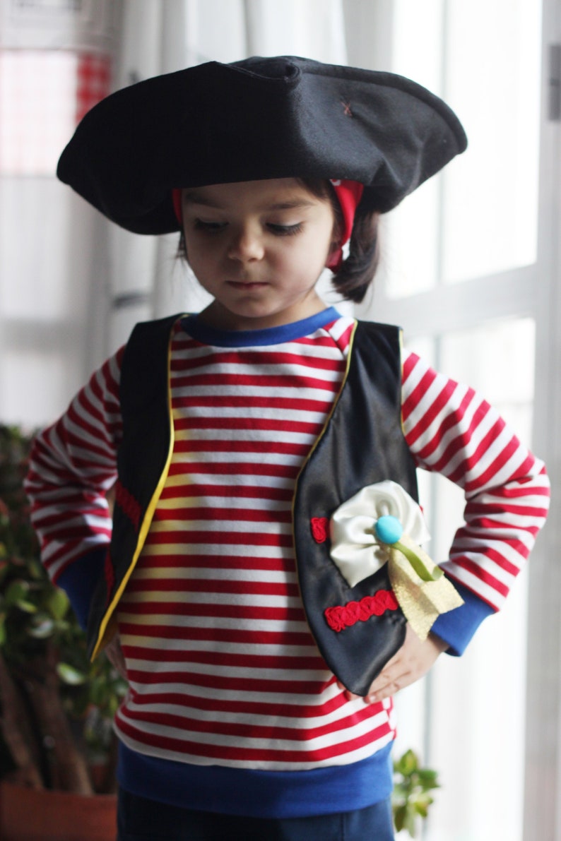May include: A child dressed in a pirate costume. The outfit includes a black tricorn hat, a red and white striped long-sleeved shirt, a black vest with gold trim, and a red bandana. The vest has a decorative white bow and a blue and gold accent.