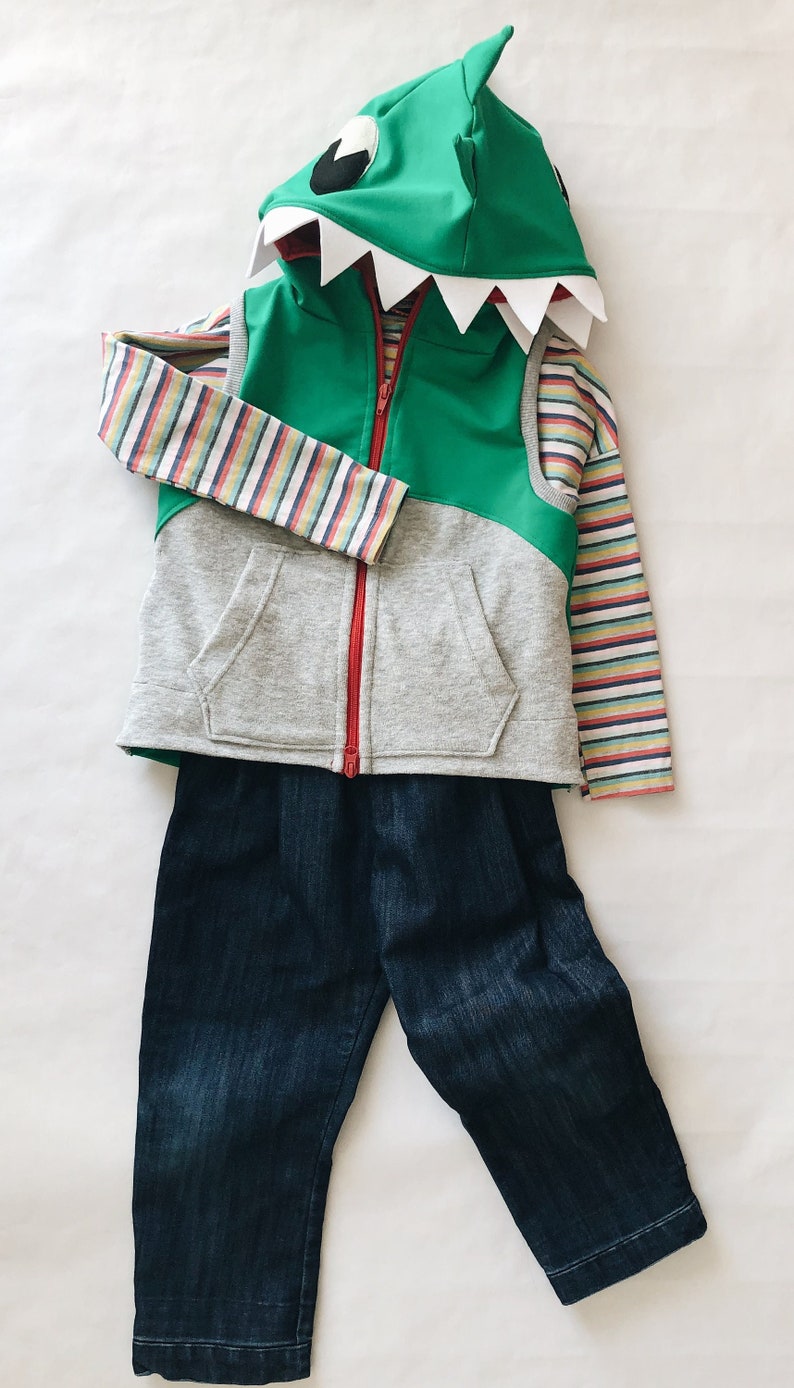 Puede incluir: Un chaleco con capucha gris y verde con una cremallera roja y una cara de tibur&oacute;n en la capucha. El chaleco se combina con una camisa de manga larga a rayas y unos vaqueros azul oscuro.