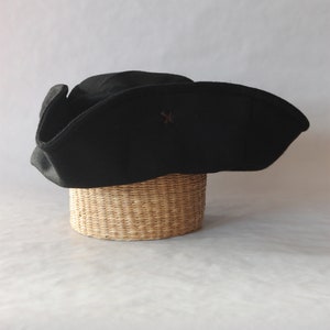 May include: A black tricorn hat with a small brown emblem, resting on a woven basket. The hat has a wide brim and a distinctive shape, often associated with historical or pirate themes. The hat is made of a dark fabric.