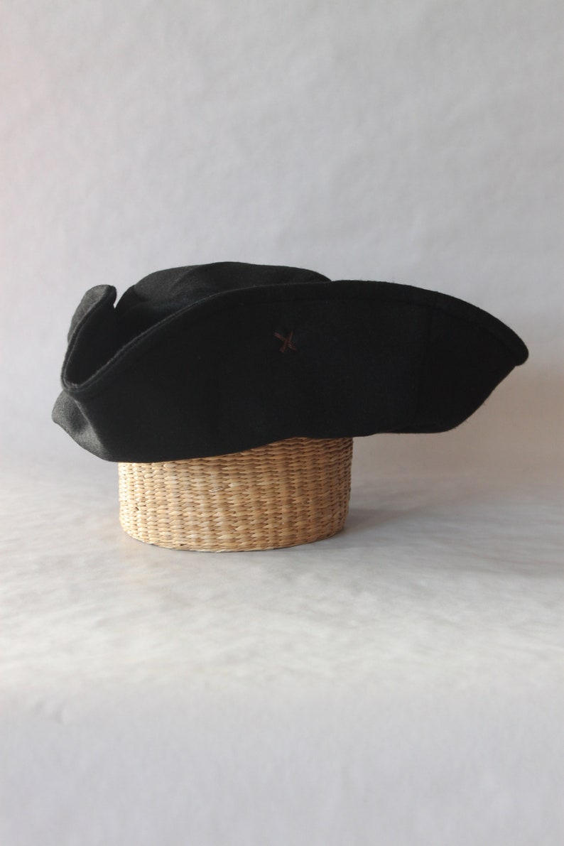 May include: A black tricorn hat with a small brown emblem, resting on a woven basket. The hat has a wide brim and a distinctive shape, often associated with historical or pirate themes. The hat is made of a dark fabric.