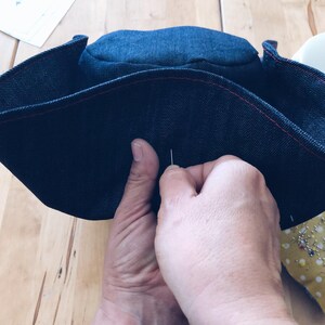Pode incluir: Uma vista de perto de um chap&eacute;u de ganga azul escuro a ser costurado &agrave; m&atilde;o. O chap&eacute;u tem costuras vermelhas. As m&atilde;os seguram uma agulha e linha. O fundo &eacute; uma superf&iacute;cie de madeira, sugerindo um projeto de costura.