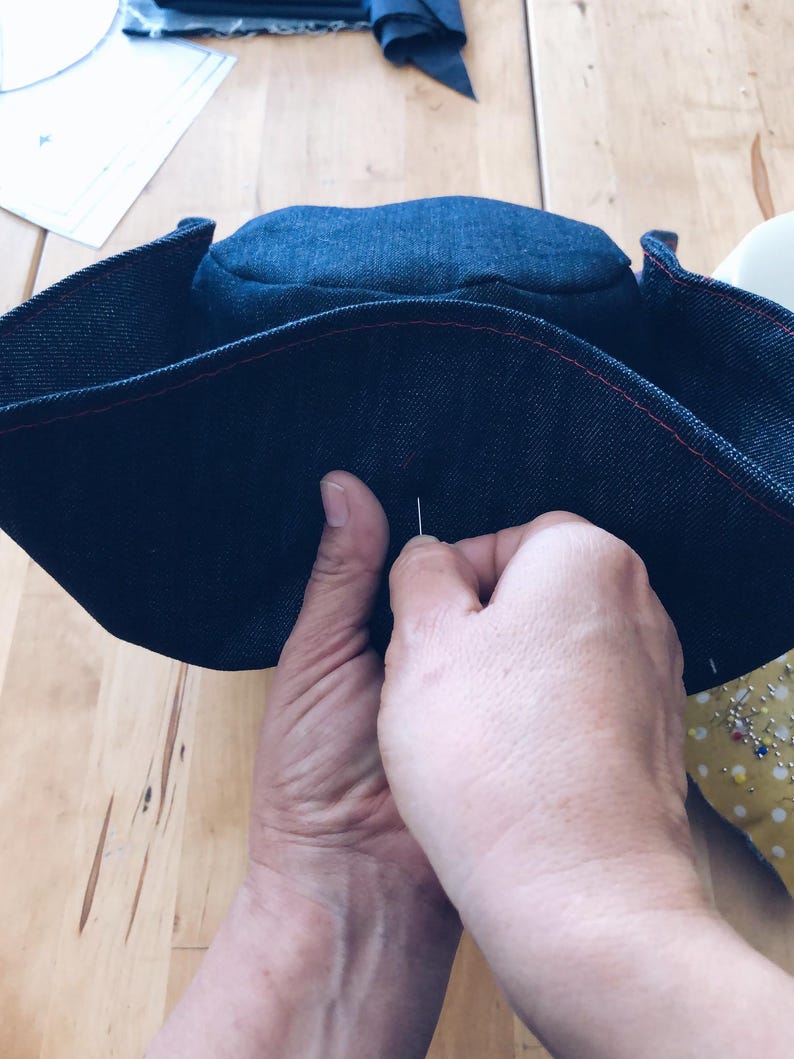 Pode incluir: Uma vista de perto de um chap&eacute;u de ganga azul escuro a ser costurado &agrave; m&atilde;o. O chap&eacute;u tem costuras vermelhas. As m&atilde;os seguram uma agulha e linha. O fundo &eacute; uma superf&iacute;cie de madeira, sugerindo um projeto de costura.