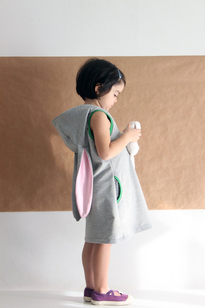 Puede incluir: Una persona joven lleva un vestido gris con capucha, orejas de conejo rosas y ribete verde. El vestido tiene un bolsillo con un dise&ntilde;o de rayas blancas y negras. La persona sostiene un animal de peluche blanco.