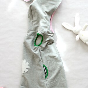 Puede incluir: Un vestido gris con capucha, orejas de conejo rosas, detalles verdes y blancos y un dise&ntilde;o de flor blanca.