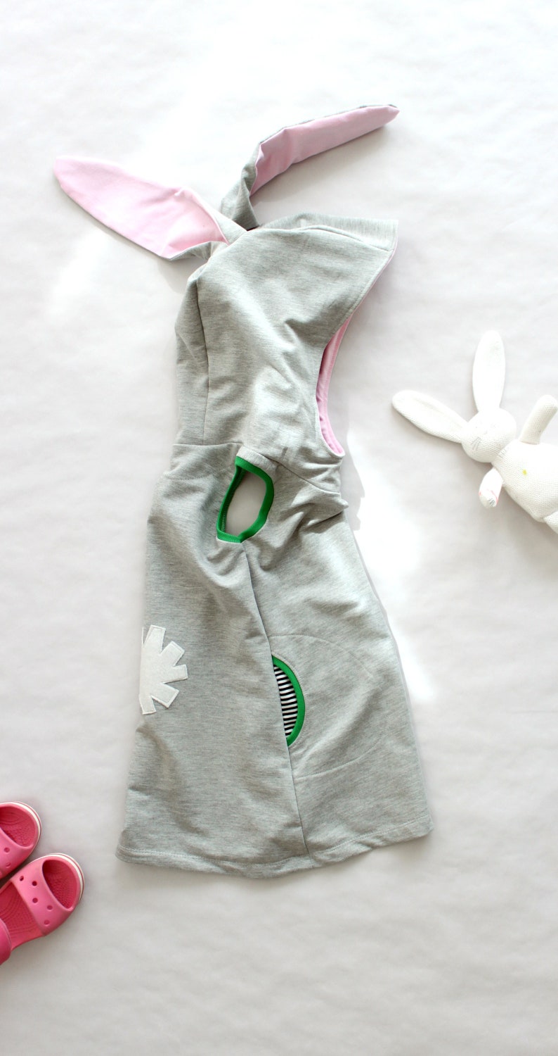 Puede incluir: Un vestido gris con capucha, orejas de conejo rosas, detalles verdes y blancos y un dise&ntilde;o de flor blanca.