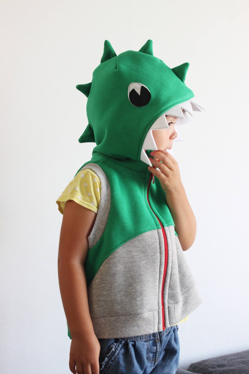 Puede incluir: Un ni&ntilde;o lleva un chaleco con capucha de dinosaurio verde y gris con una cremallera blanca. La capucha tiene una boca blanca con dientes y un ojo negro.