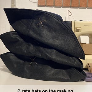 May include: Three black pirate hats stacked on top of each other. Each hat has a gold-colored cross stitched on the front. The text "Pirate hats on the making. Here in color black." is at the bottom of the image.