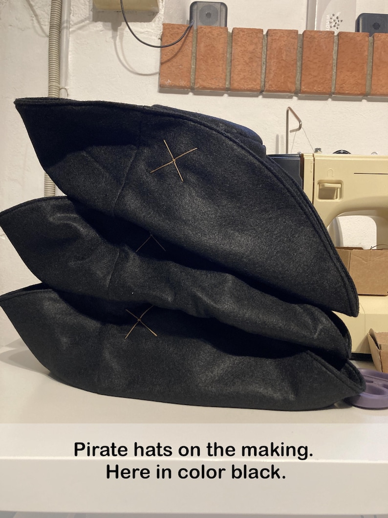 May include: Three black pirate hats stacked on top of each other. Each hat has a gold-colored cross stitched on the front. The text "Pirate hats on the making. Here in color black." is at the bottom of the image.
