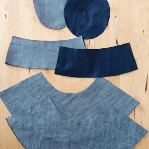 Pode incluir: V&aacute;rias pe&ccedil;as de tecido de ganga em diferentes formas, incluindo dois c&iacute;rculos, dois ret&acirc;ngulos curvos e uma pe&ccedil;a maior e curva. Os tecidos s&atilde;o tons de azul, dispostos sobre uma superf&iacute;cie castanha clara.