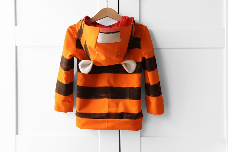 May include: Orange and brown striped hooded sweatshirt with attached ears. The long-sleeved top is hanging on a wooden hanger. The hood has a white and tan patch.