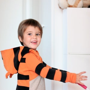 May include: A child wearing an orange and black striped hooded sweatshirt, resembling a tiger. The child is smiling and holding a pink and white object. The background includes a white door and a white cabinet.