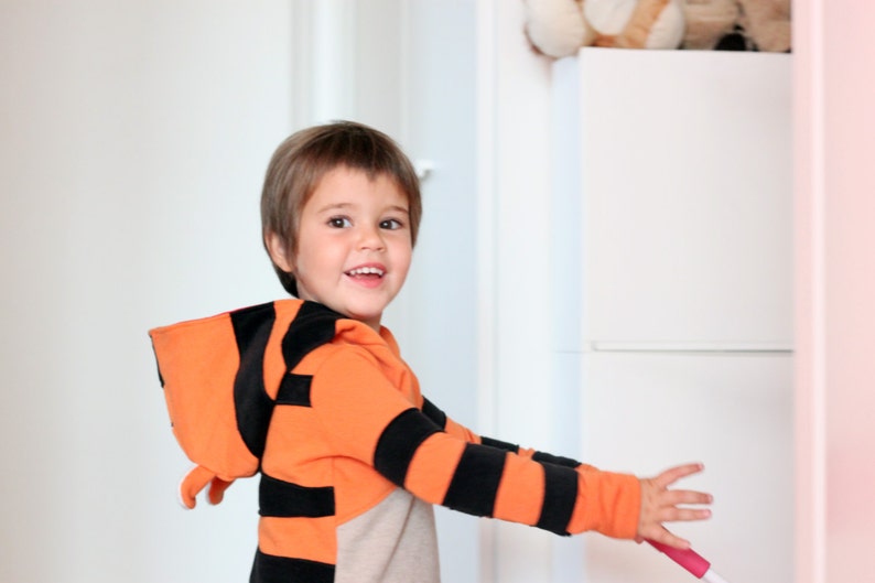 May include: A child wearing an orange and black striped hooded sweatshirt, resembling a tiger. The child is smiling and holding a pink and white object. The background includes a white door and a white cabinet.