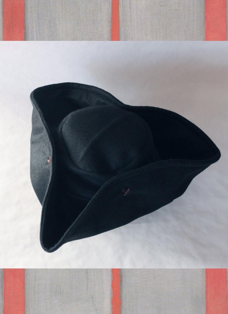 May include: A black tricorn hat with a rounded crown and upturned brims. The hat features small, decorative buttons and is set against a white background with a textured, gray and red border.