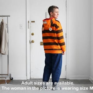 May include: An orange and brown striped hooded sweatshirt with a red interior and white ear-shaped accents. The person in the image is wearing blue jeans. The text on the image says "Adult sizes are available. The woman in the picture is wearing size M."