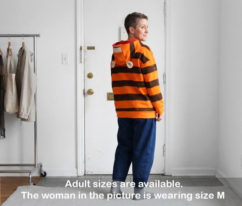 May include: An orange and brown striped hooded sweatshirt with a red interior and white ear-shaped accents. The person in the image is wearing blue jeans. The text on the image says "Adult sizes are available. The woman in the picture is wearing size M."