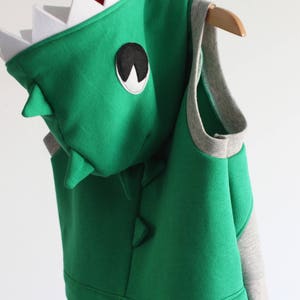 Puede incluir: Chaleco con capucha de dinosaurio verde con ribete gris y parche negro para el ojo. La capucha tiene dientes blancos.