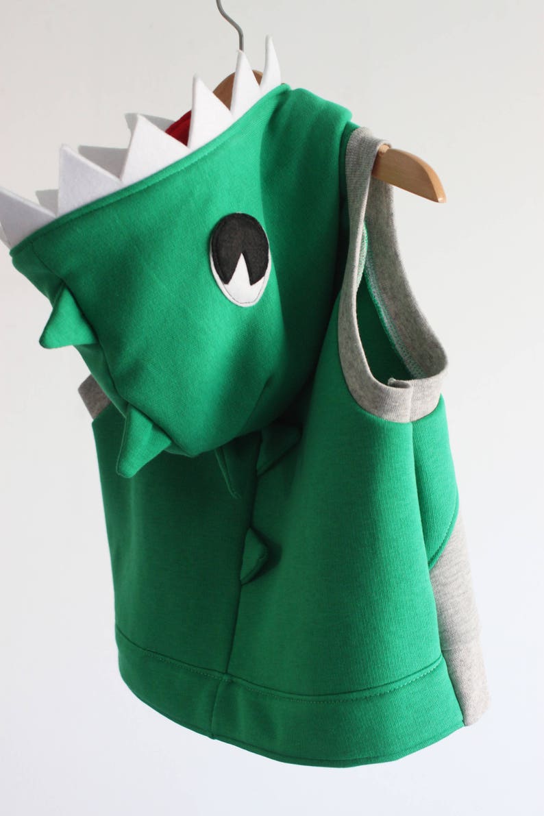 Puede incluir: Chaleco con capucha de dinosaurio verde con ribete gris y parche negro para el ojo. La capucha tiene dientes blancos.