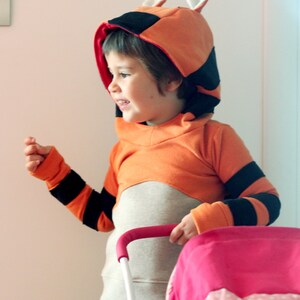 May include: An orange and black hooded sweatshirt with a tiger design. The sweatshirt has black stripes on the sleeves and a hood with ears. The child is wearing blue jeans and holding a pink stroller.
