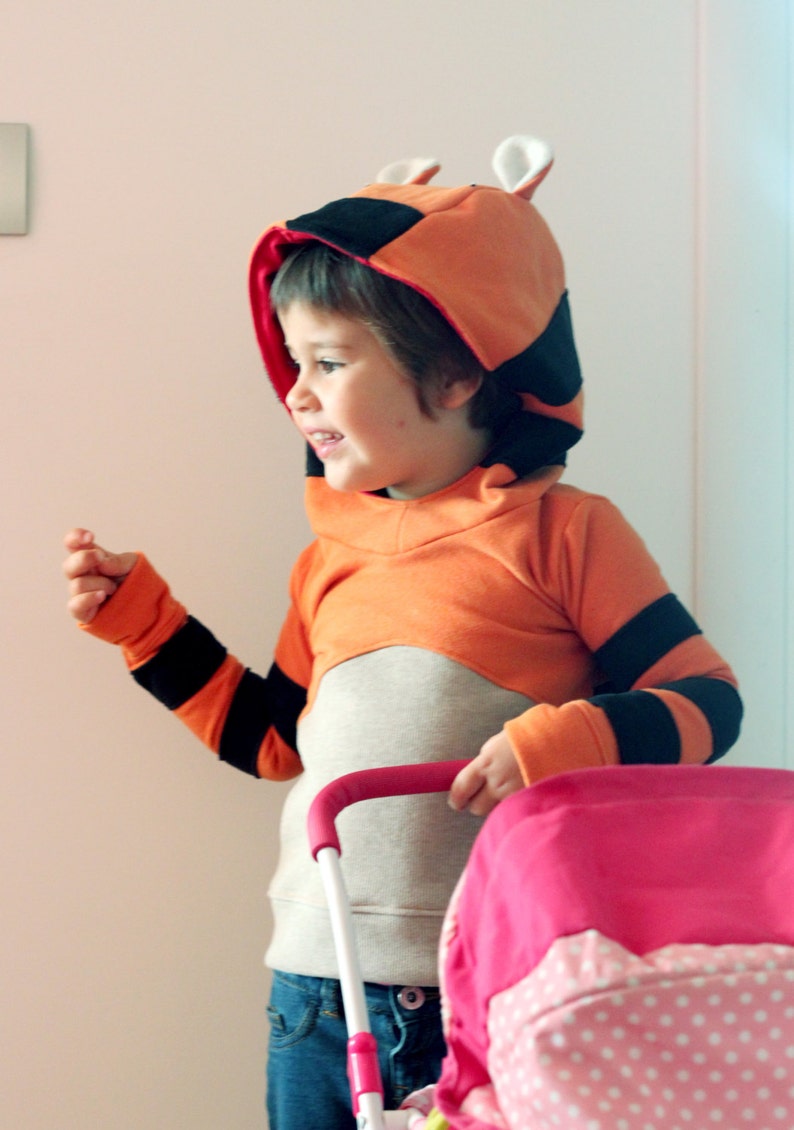 May include: An orange and black hooded sweatshirt with a tiger design. The sweatshirt has black stripes on the sleeves and a hood with ears. The child is wearing blue jeans and holding a pink stroller.