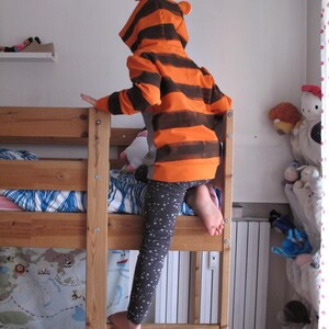May include: A child wearing an orange and brown striped tiger hoodie with matching ears, climbing a wooden bunk bed. The child is also wearing gray leggings with white star patterns. The bed has a map-themed bedspread.
