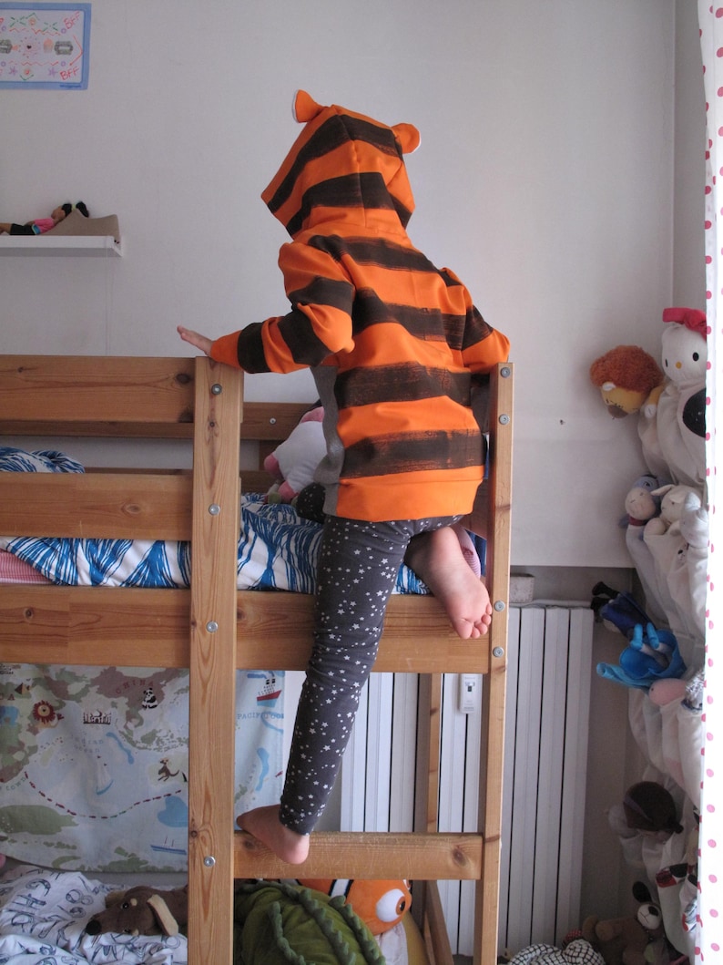 May include: A child wearing an orange and brown striped tiger hoodie with matching ears, climbing a wooden bunk bed. The child is also wearing gray leggings with white star patterns. The bed has a map-themed bedspread.