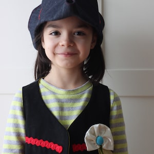 Pode incluir: Uma crian&ccedil;a usando um chap&eacute;u de ganga azul escuro com costuras vermelhas e um colete preto com acabamentos vermelhos. O colete tem uma flor branca com centro azul e fita verde. A crian&ccedil;a est&aacute; vestindo uma camisa listrada.