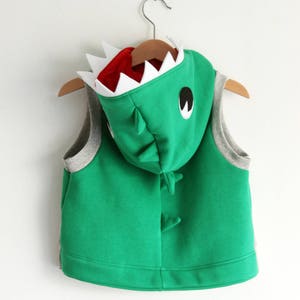 Puede incluir: Chaleco verde con capucha y dise&ntilde;o de dinosaurio. La capucha tiene una boca blanca y roja con dientes y un ojo negro.