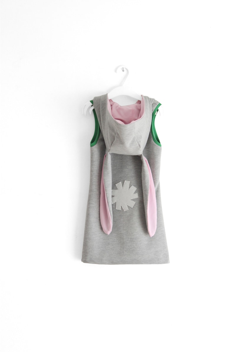 Puede incluir: Un vestido gris con capucha, orejas de conejo rosas y una flor gris en la espalda. El vestido tiene un ribete verde alrededor del cuello y las sisas.