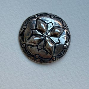 May include: A circular silver-toned metal button featuring a central four-pointed star design composed of raised diamond shapes. The outer edge is decorated with a series of small circular indentations and fine radial lines etched into the metal.