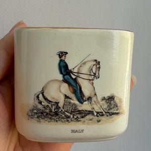 May include: A cream-colored ceramic mug with a gold-toned rim. The front features a detailed illustration of a rider in a blue jacket on a white horse, with the word HALT printed in black capital letters below the scene.