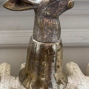 May include: A metallic bell featuring a sculpted hare head handle with detailed fur texture and long ears. The bell body has a tarnished brass finish with visible patina and dark spots, showing a flared base and a smooth, rounded top section.