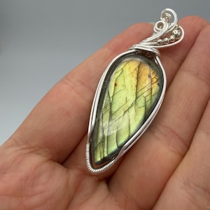 May include: A teardrop-shaped labradorite pendant featuring a polished, iridescent green and gold stone with visible internal fractures. The gemstone is encased in a decorative silver wire-wrapped setting with intricate coils and small metal beads.
