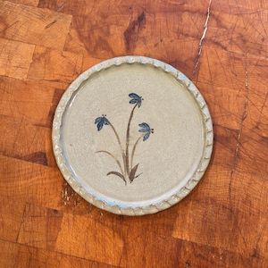May include: A round ceramic plate with a beige speckled surface and a scalloped edge. The center features a hand-painted design of three blue flowers with brown stems and leaves. The plate has a smooth, glazed finish and a neutral color palette.