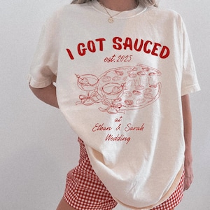 May include: Cream-colored t-shirt with a red graphic that reads "I GOT SAUCED est. 2025" above a pizza and two cocktails. Below the graphic is "at Ethan & Sarah Wedding".